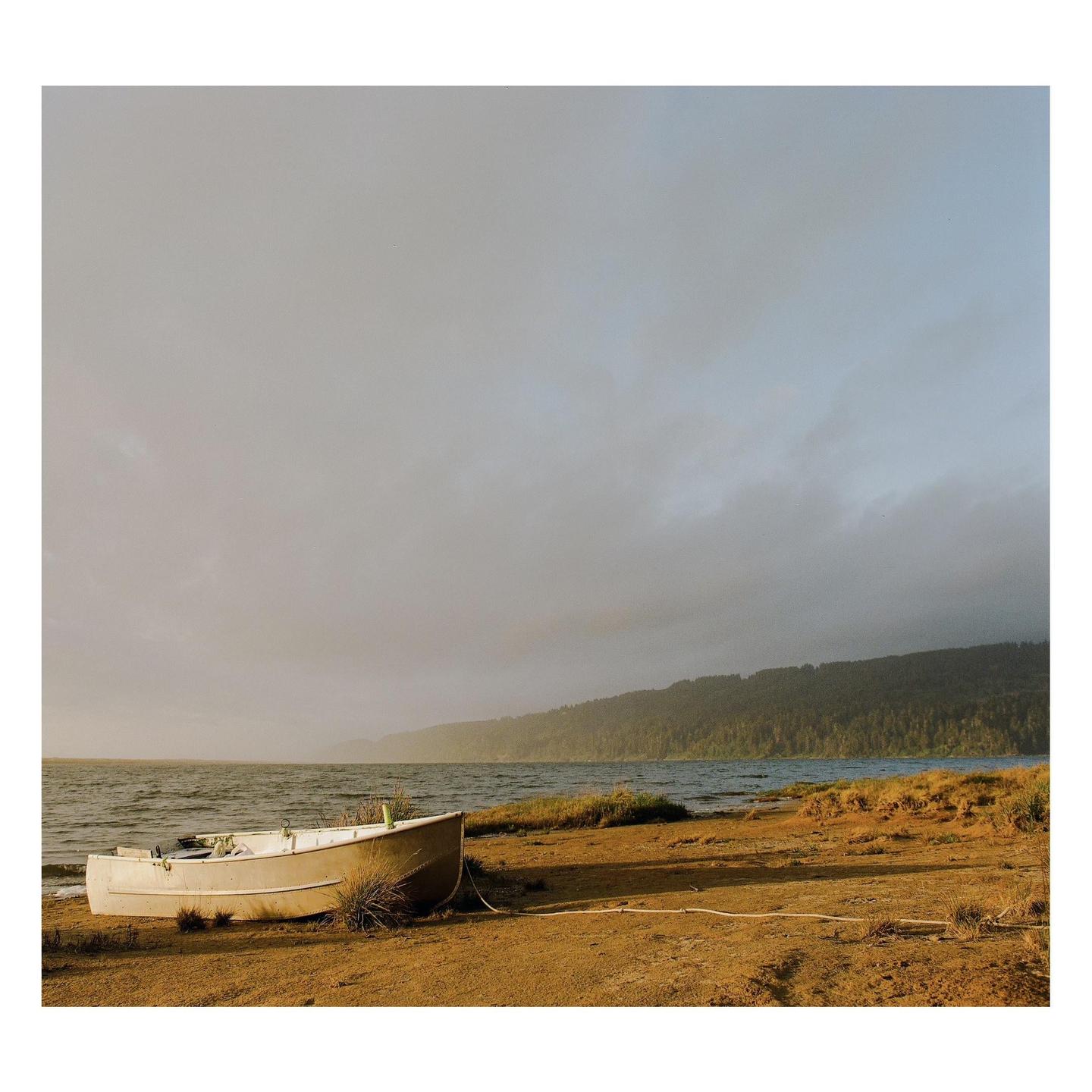 A small boat rests on a sandy shore next to a body of water with a forested hill in the distance under a cloudy sky.