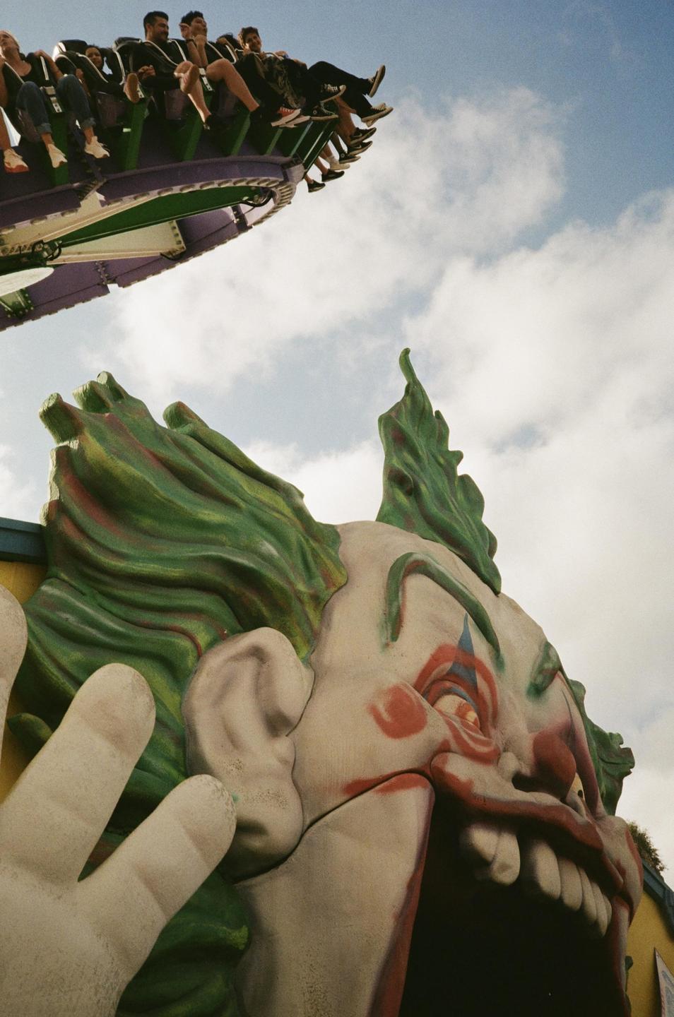 A group of people are riding a roller coaster above a large clown head entrance at an amusement park.