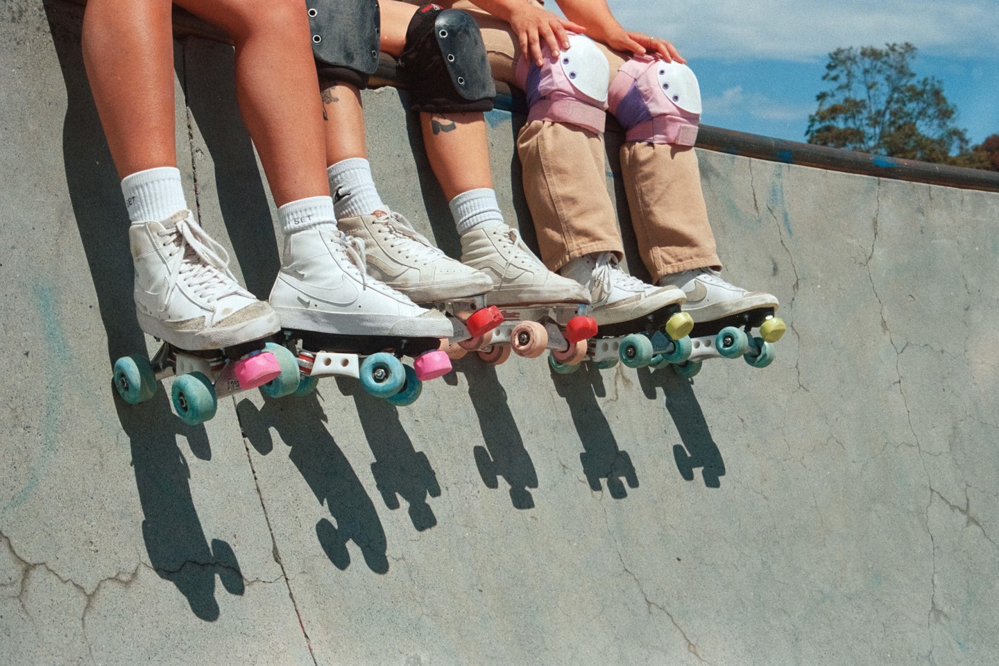 Four people sit on the edge of a skate park wearing roller skates and knee pads.