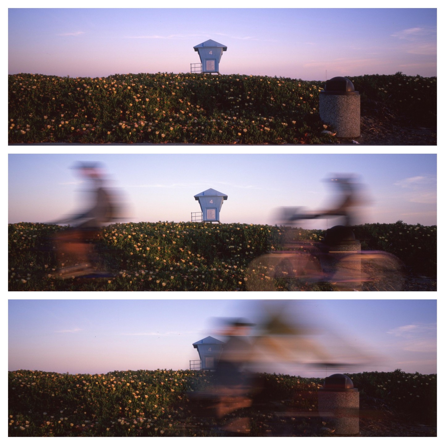 A blue lifeguard stand sits atop a grassy hill covered in small yellow flowers, with blurred figures of people on bicycles passing by in the foreground.