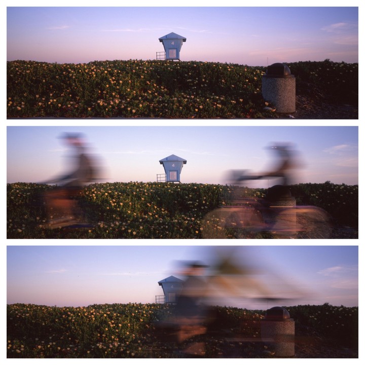 A blue lifeguard stand sits atop a grassy hill covered in small yellow flowers, with blurred figures of people on bicycles passing by in the foreground.