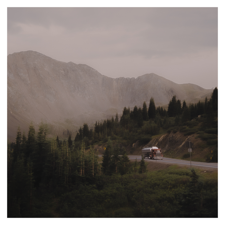 A truck drives along a winding road through a mountainous landscape with pine trees under a cloudy sky.
