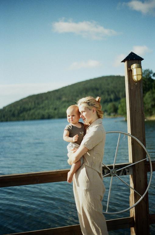 A woman holds a baby on a wooden dock next to a body of water with a forested hill in the background.