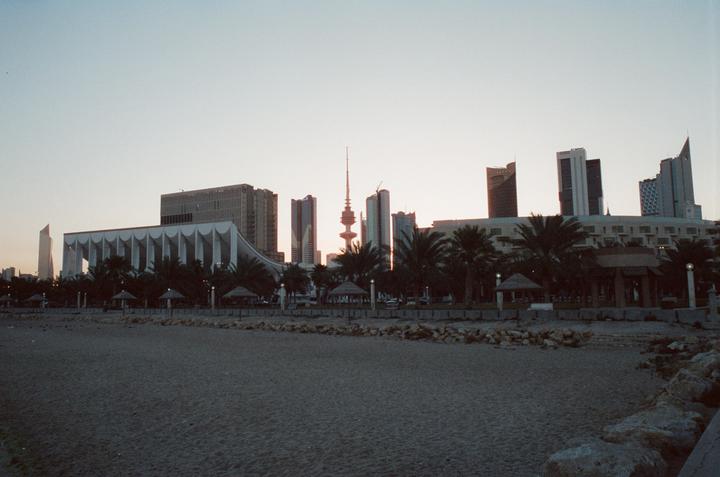 A sandy beach with rocks in the foreground looks towards a city skyline with palm trees and a prominent tower at dusk.