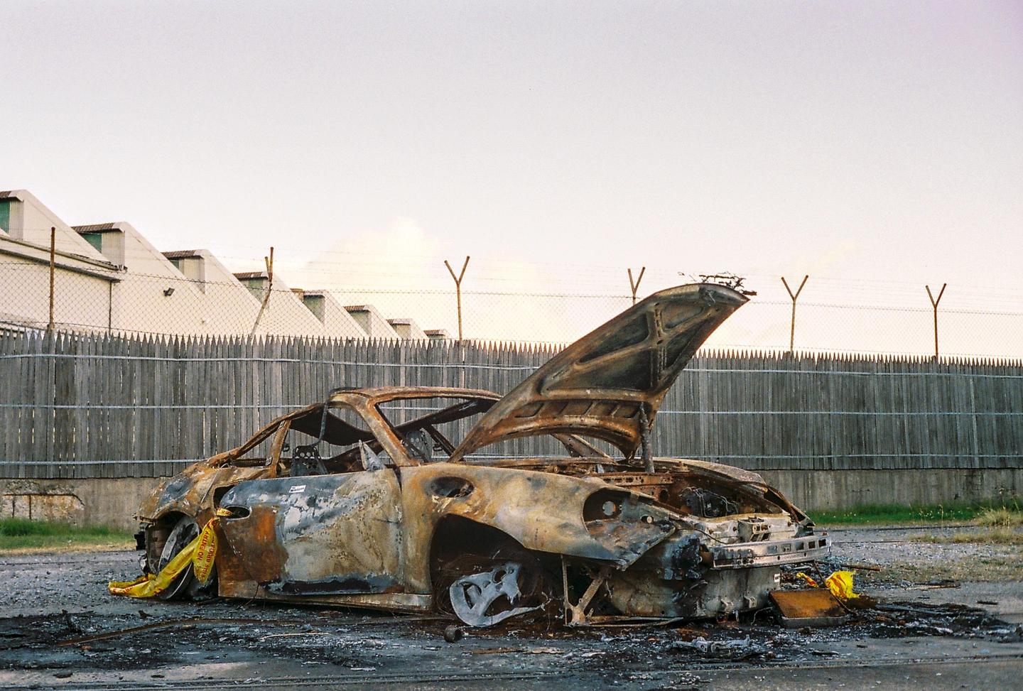 A burnt-out car with its hood open sits on an asphalt lot next to a wooden fence and a building.