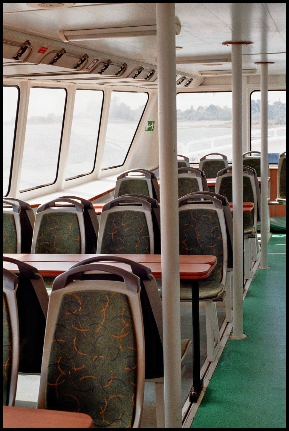 The interior of a ferry shows rows of empty seats and tables next to large windows looking out onto water and land.