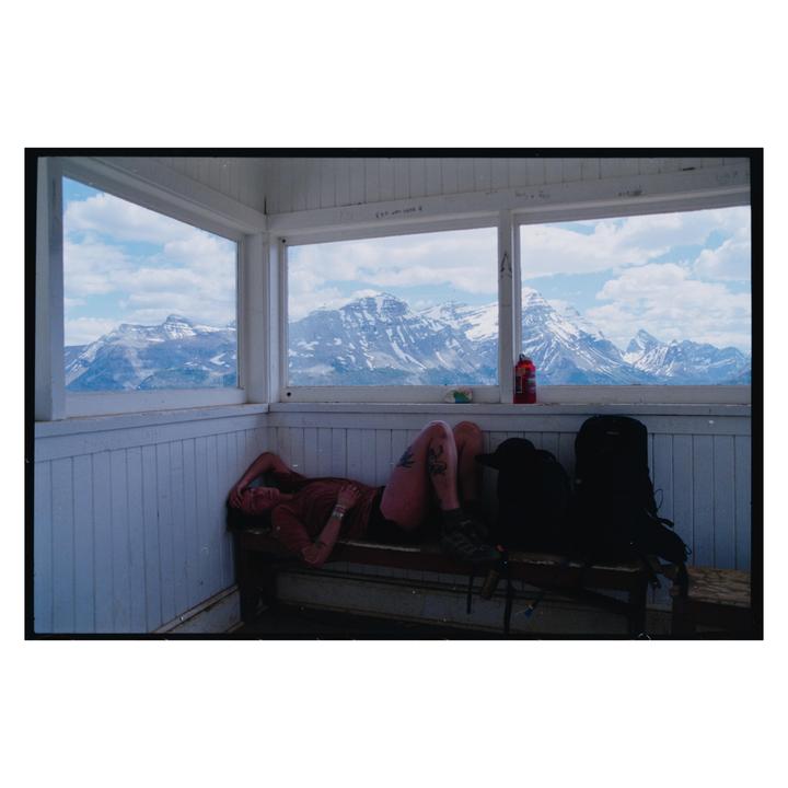 A woman rests on a bench inside a building with large windows overlooking snow-capped mountains.