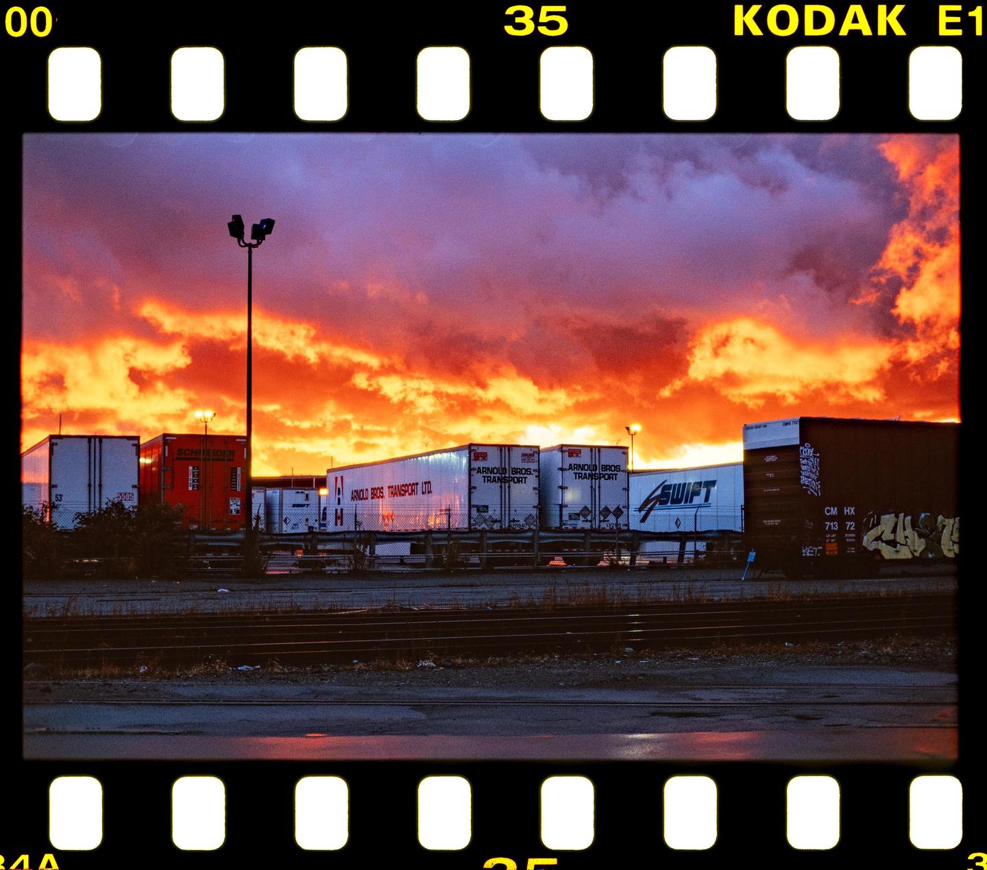 A dramatic sunset with orange and red clouds illuminates a row of parked semi-trailers and a street light.