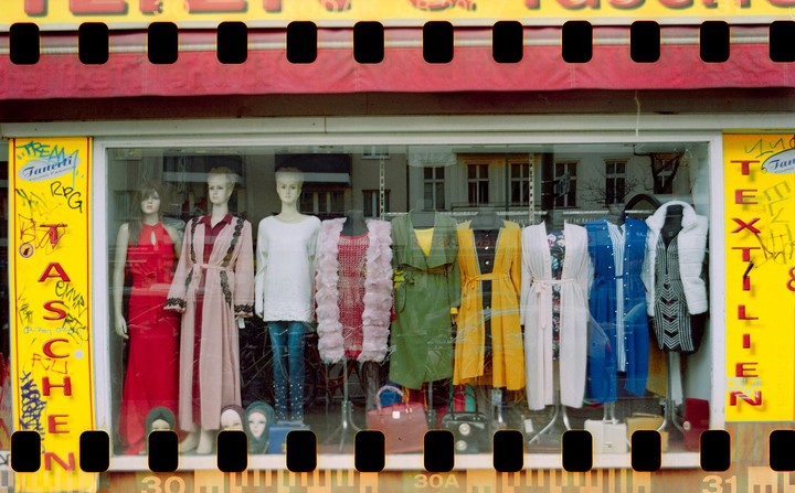 Mannequins display colorful clothing in a shop window with reflections of buildings and graffiti on the glass.