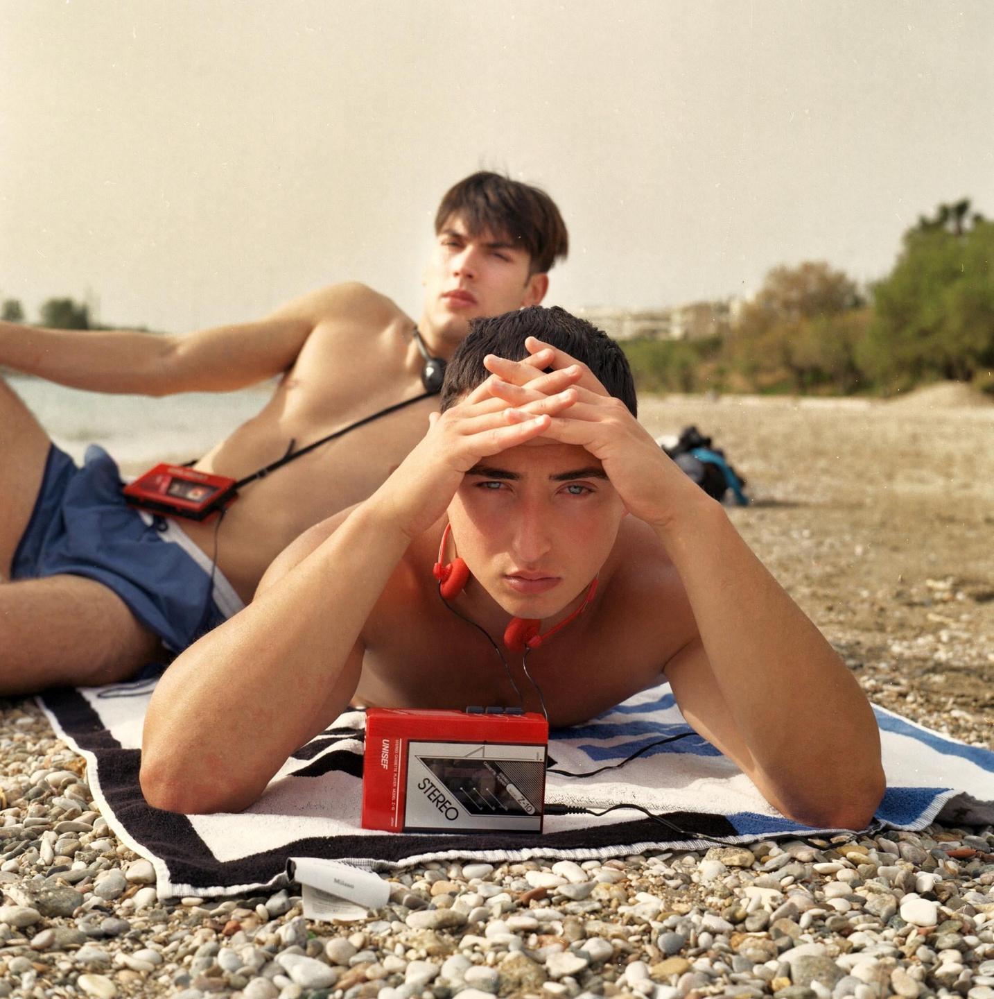 Two boys are lying on a rocky beach with a red stereo cassette player and headphones.