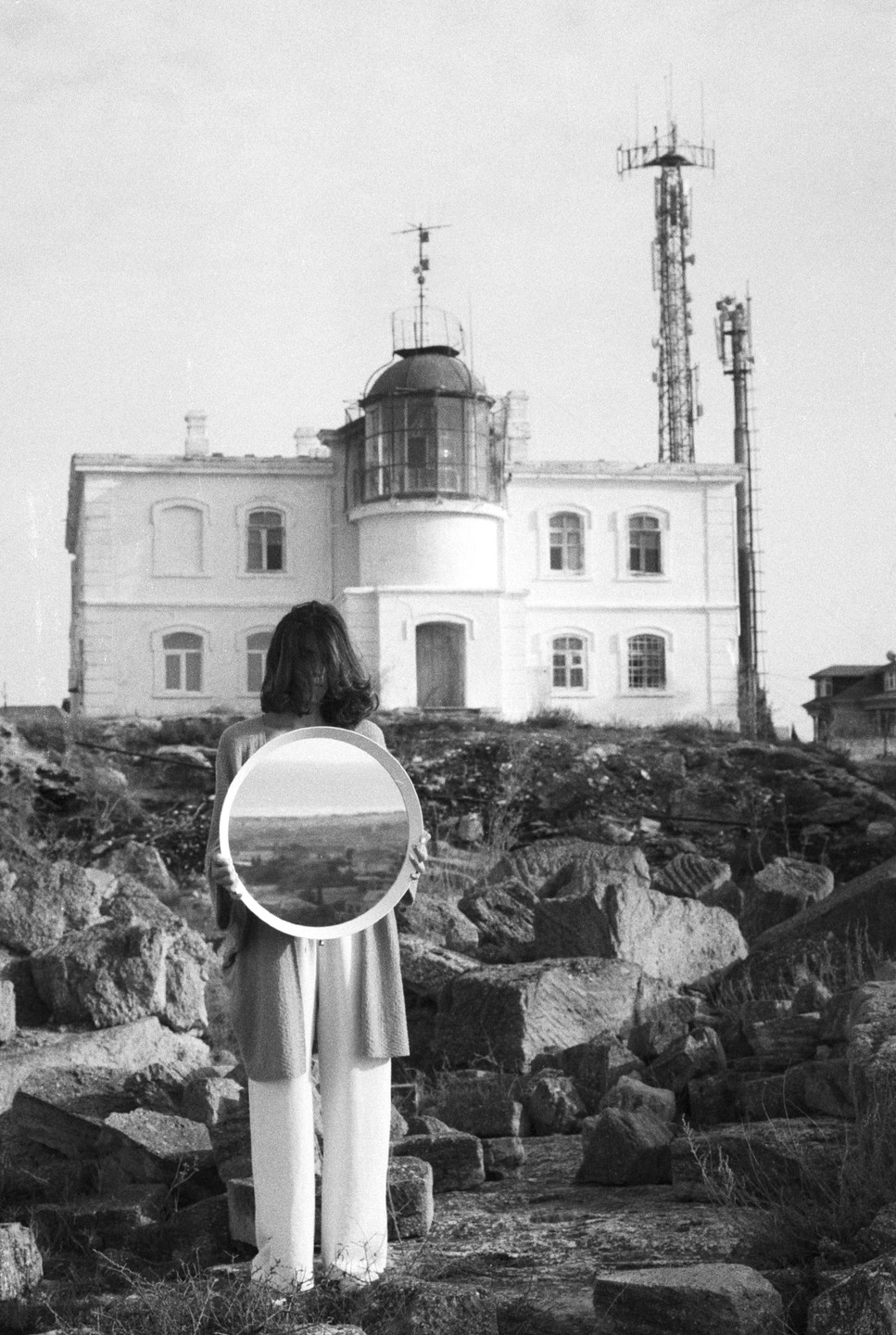 A woman stands with her back to the viewer, holding a round mirror that reflects a landscape, in front of a lighthouse and communication towers.