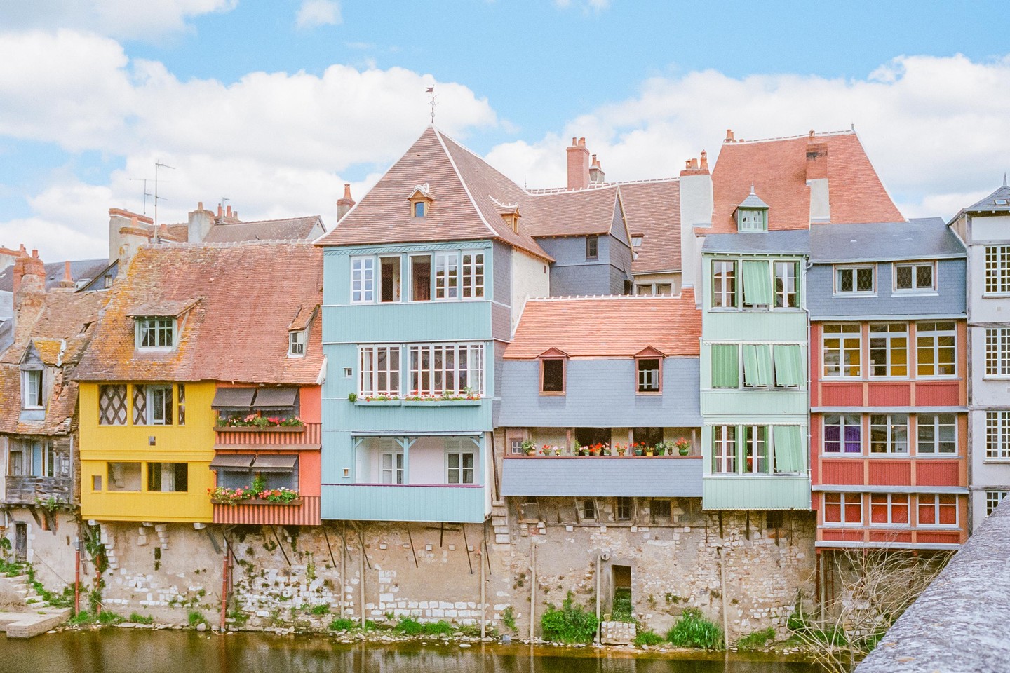 A row of colorful houses with red roofs stands along a river under a blue sky with white clouds.