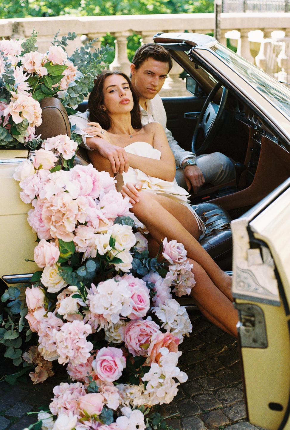 A couple sits in a convertible car overflowing with pink and white flowers.
