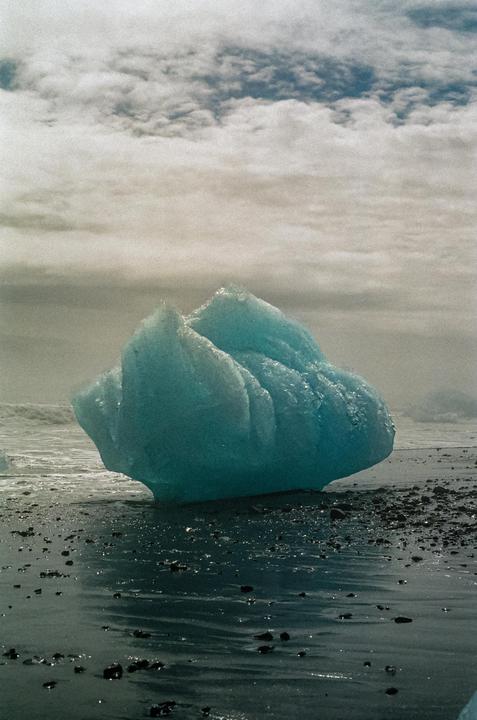A large blue iceberg rests on a black sand beach with waves in the background under a cloudy sky.