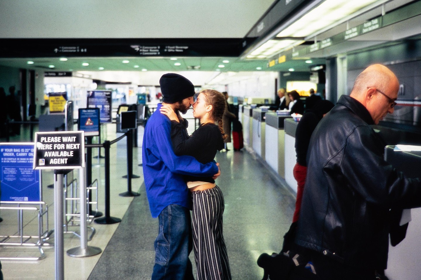A couple embraces and kisses in an airport terminal while a man checks in at a counter.