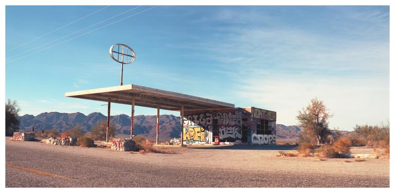 An abandoned gas station with graffiti on its walls stands in a desert landscape with mountains in the background.