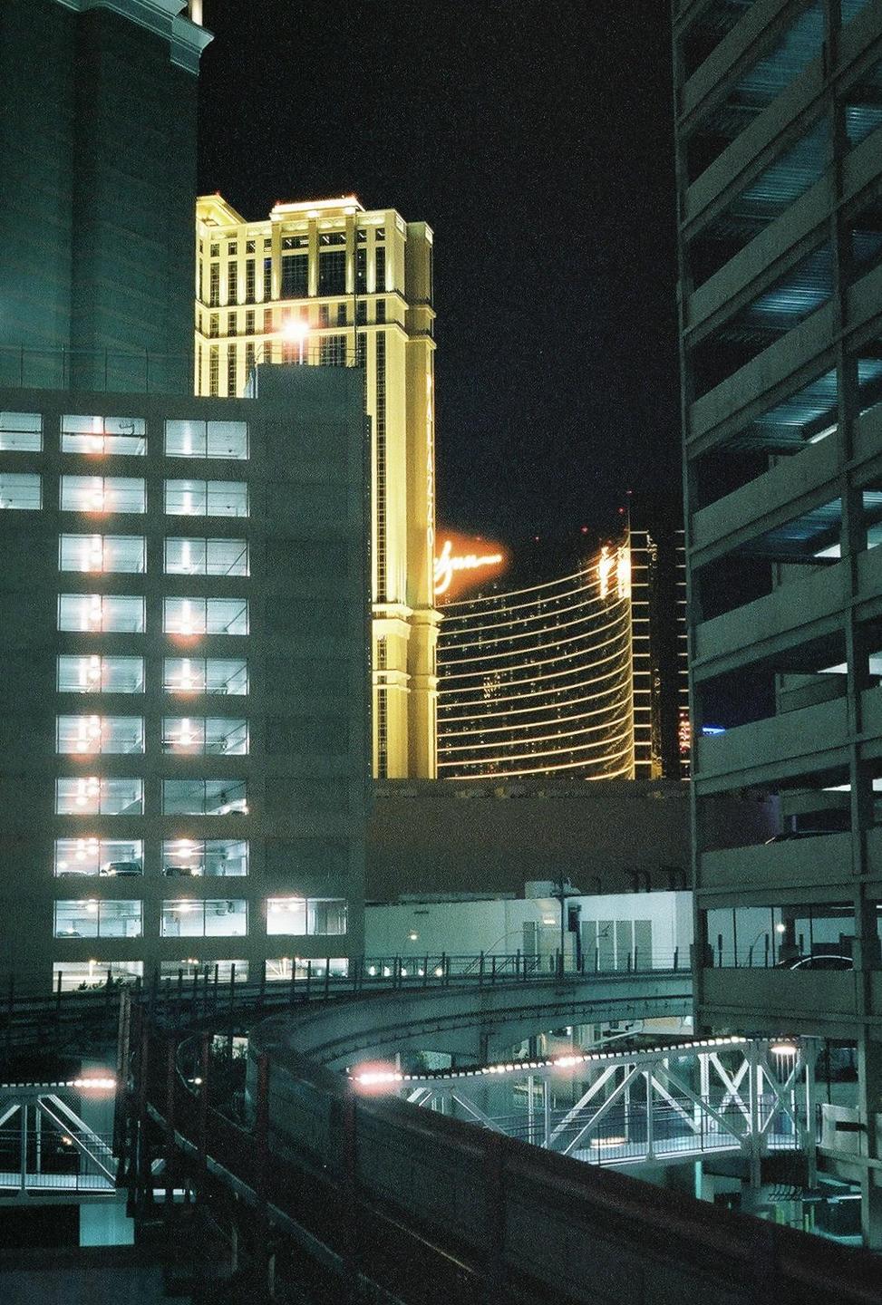 A monorail track curves through a city at night, with brightly lit buildings and a parking garage visible.