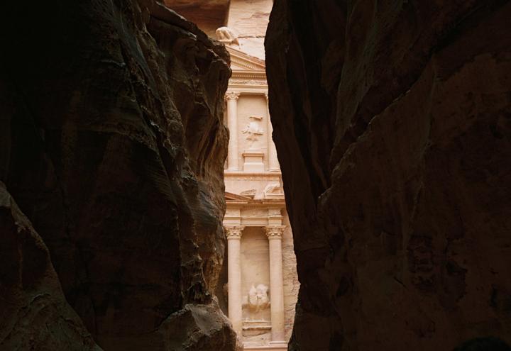 A narrow view of a carved stone building is framed by dark rock walls.