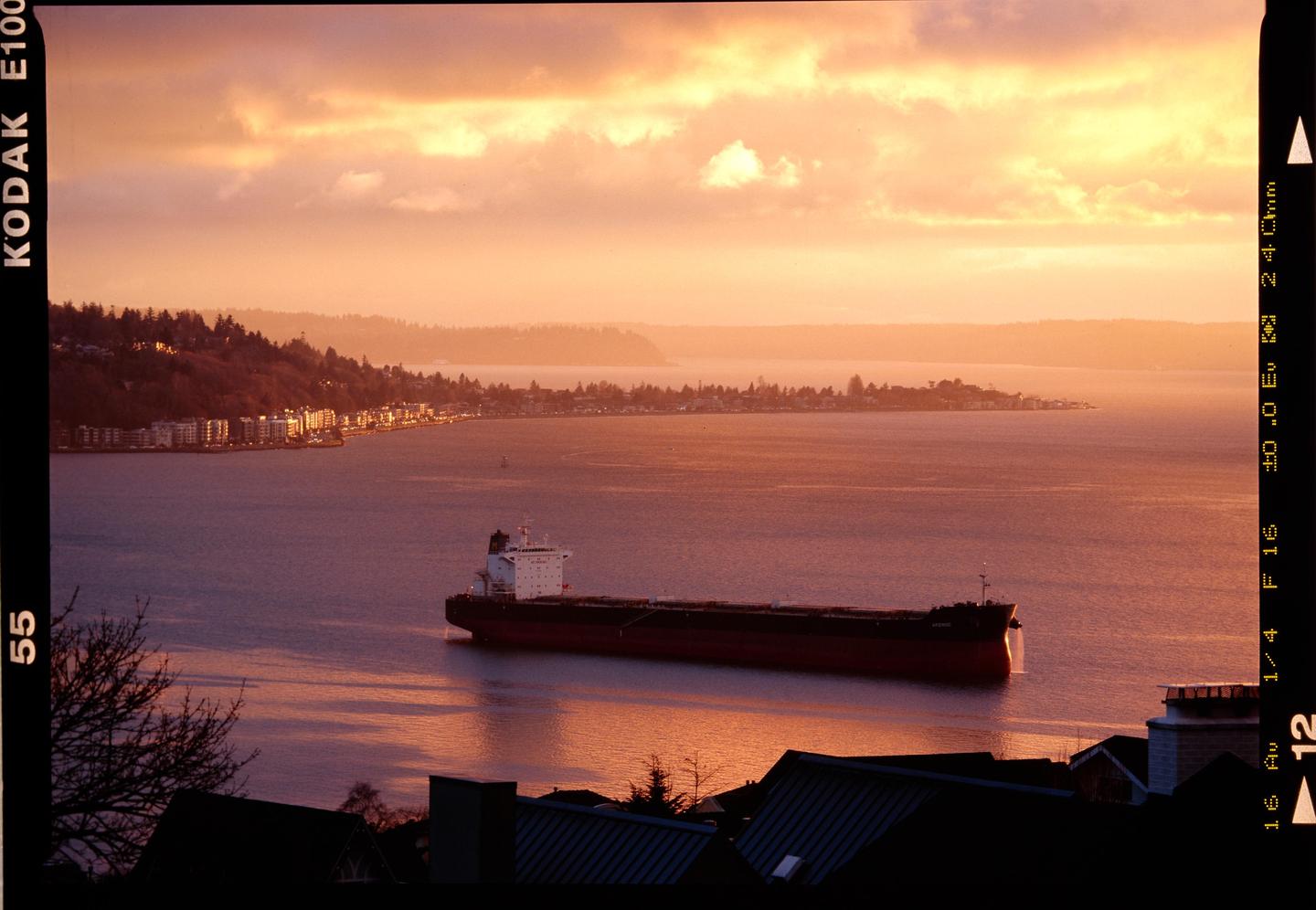 A large cargo ship floats on the water with a city and hills in the background under a dramatic sunset sky.