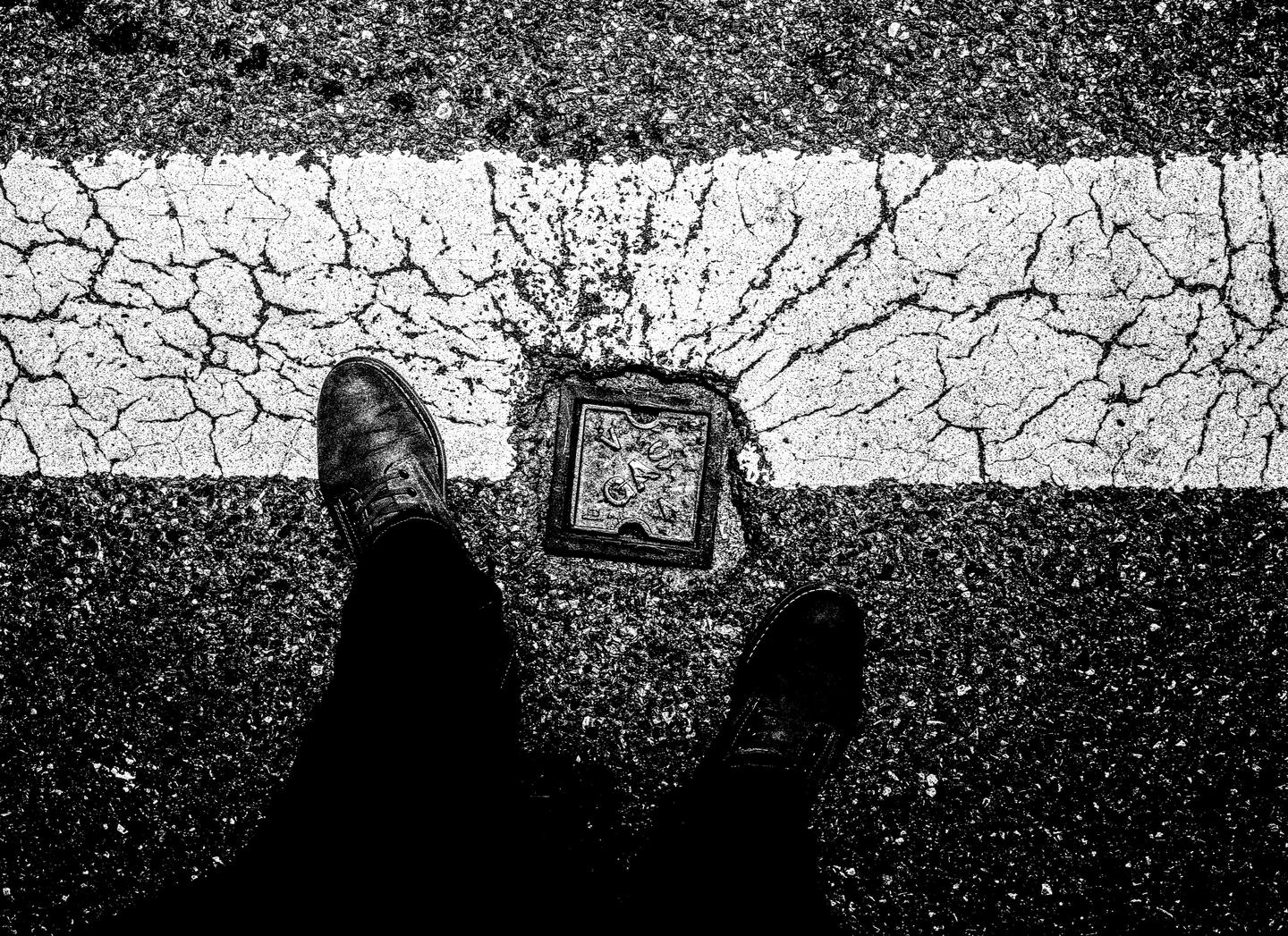 A pair of feet stand on a cracked asphalt road with a white line and a metal plate.
