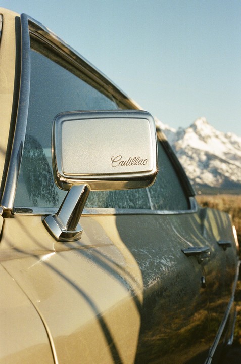 A Cadillac side mirror reflects a snowy mountain peak under a clear sky.