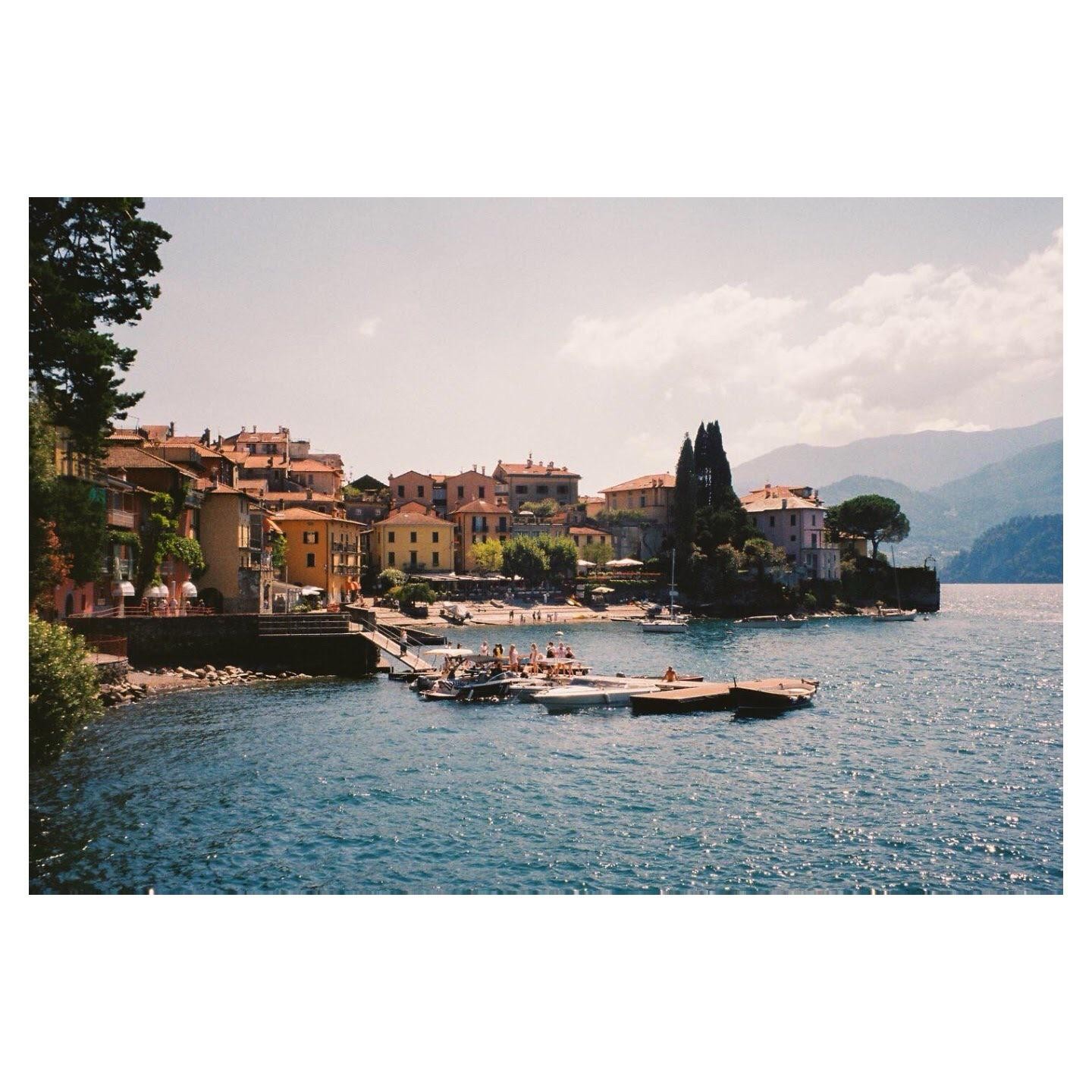 A small town with colorful buildings sits on the shore of a lake with mountains in the background.