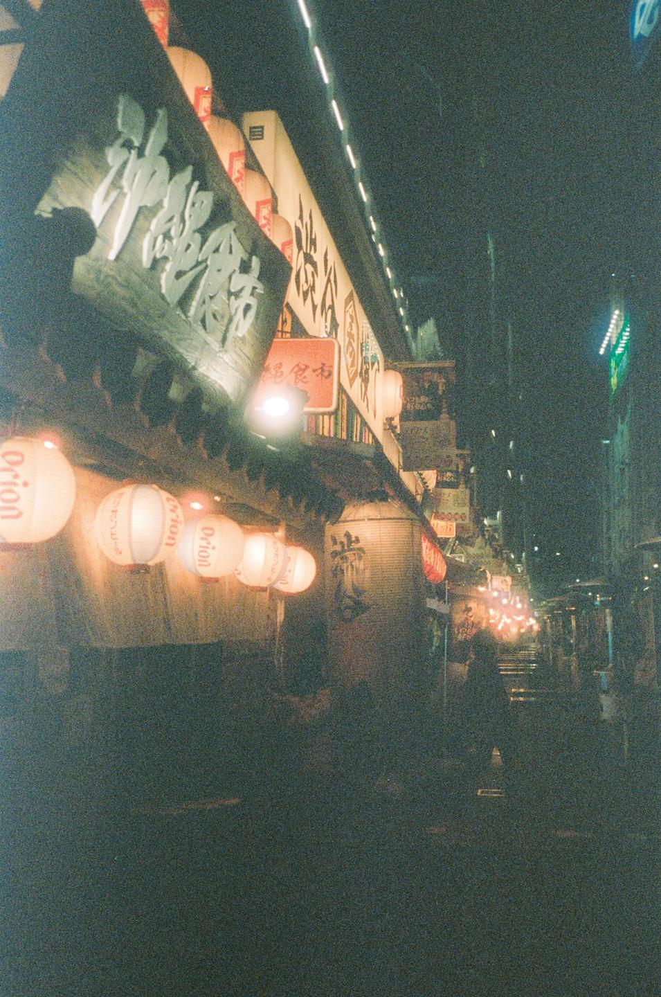 A street at night is illuminated by hanging lanterns and neon signs.
