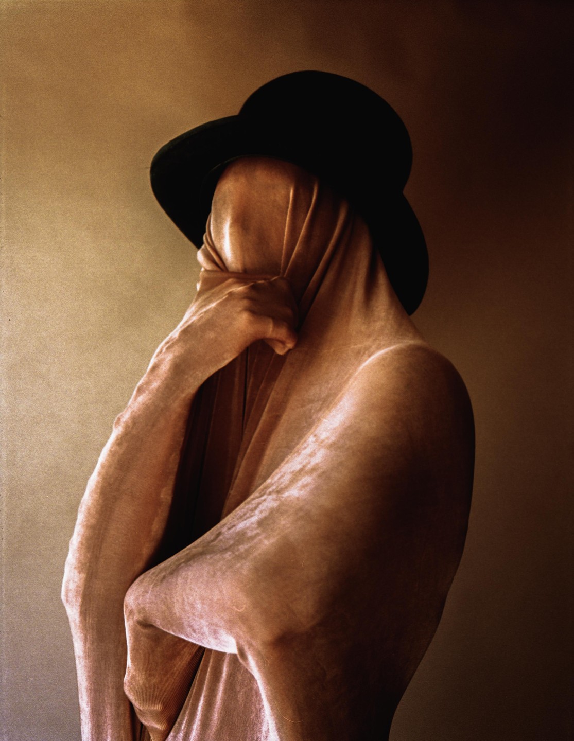 A person wearing a hat and covered in a mesh fabric holds their face with one hand.