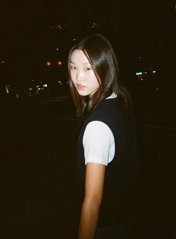 A young woman with long hair looks over her shoulder at night with city lights in the background.