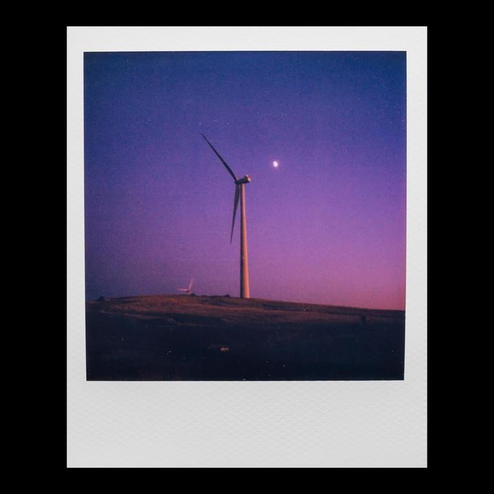 A wind turbine stands on a hill under a purple and blue sky with a crescent moon.