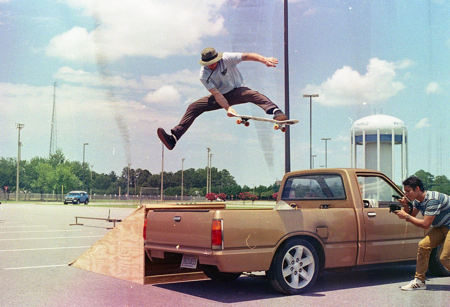 A man on a skateboard jumps over a brown pickup truck while another man photographs him in a parking lot.
