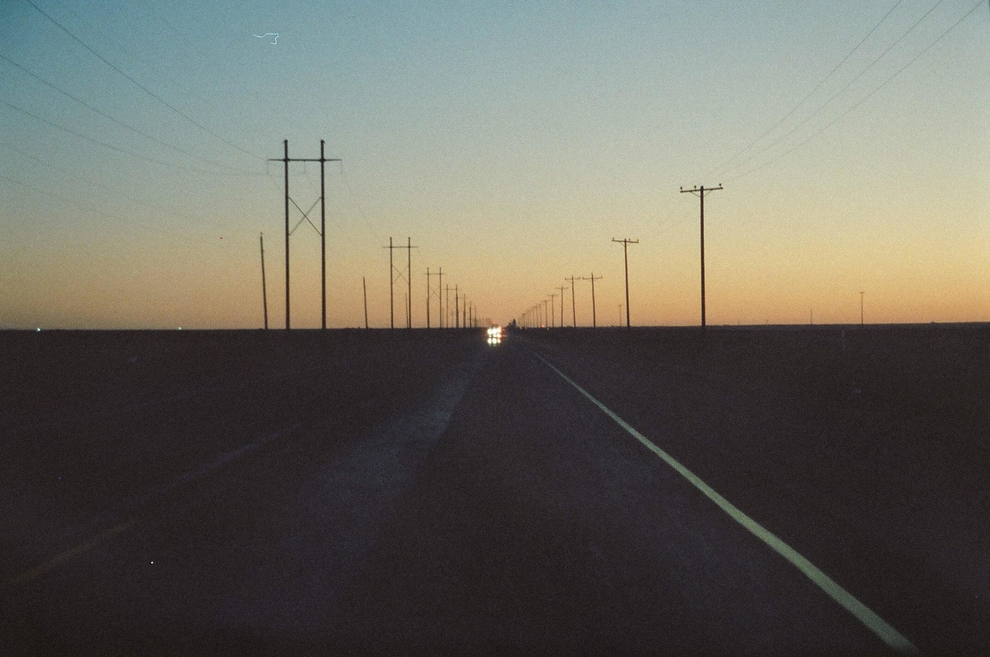 A long, straight road stretches into the distance, lined with utility poles and a car approaching with headlights on under a twilight sky.