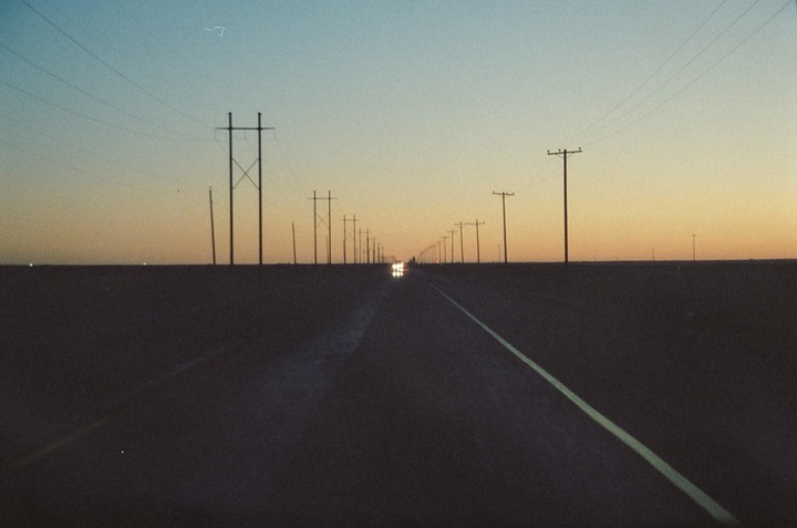 A long, straight road stretches into the distance, lined with utility poles and a car approaching with headlights on under a twilight sky.