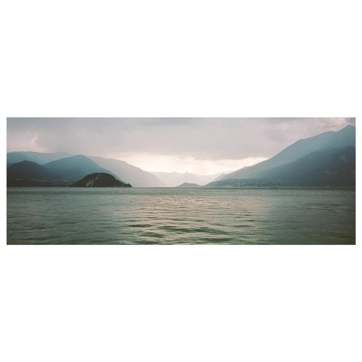 A panoramic landscape shows a lake surrounded by mountains under a cloudy sky.