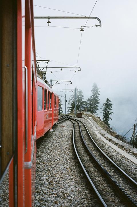 A red train is on a track next to a mountain with trees and fog.
