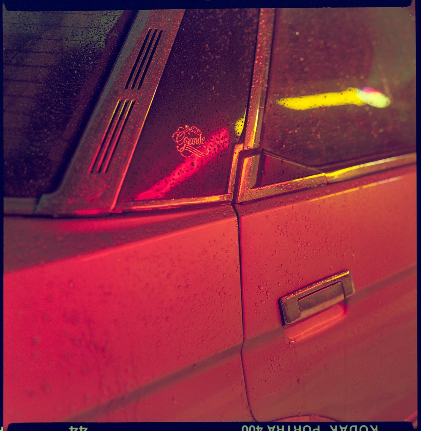 A red car is covered in raindrops, reflecting neon lights.