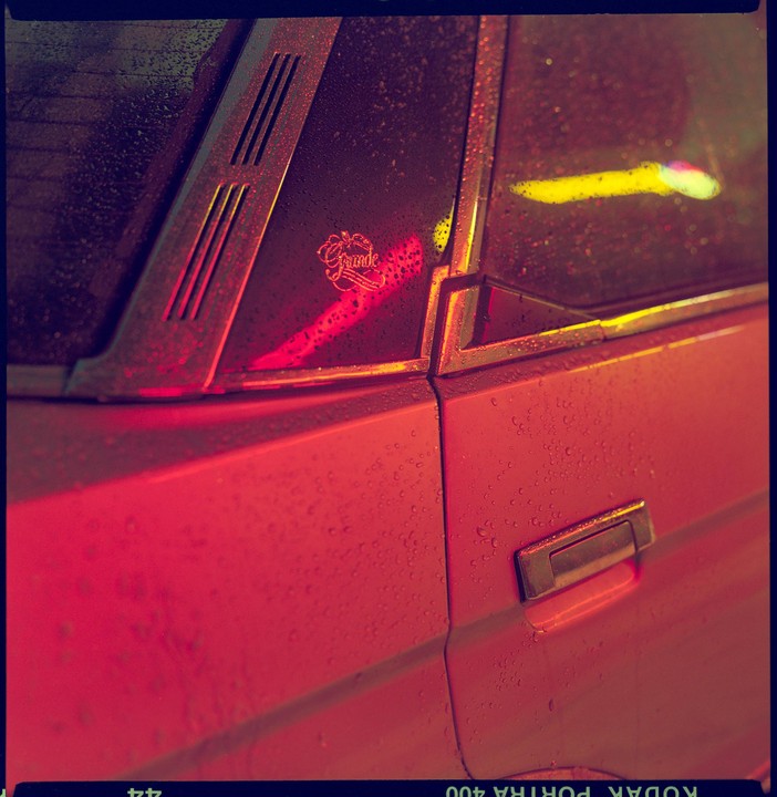 A red car is covered in raindrops, reflecting neon lights.