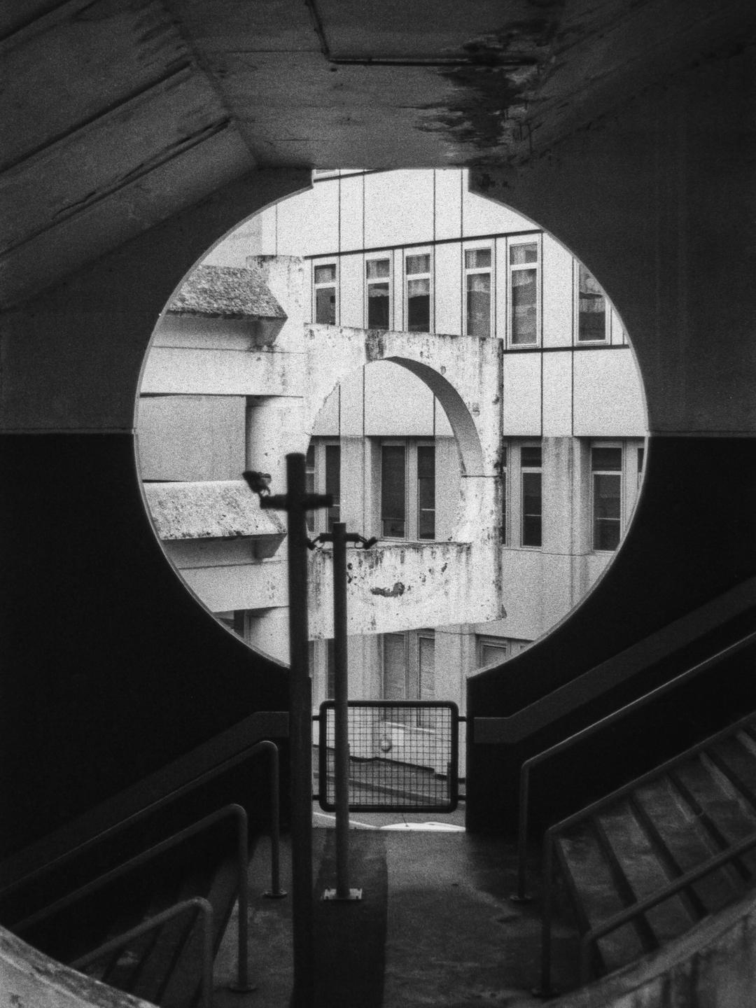 A circular opening in a concrete structure frames a building with multiple windows and an archway.