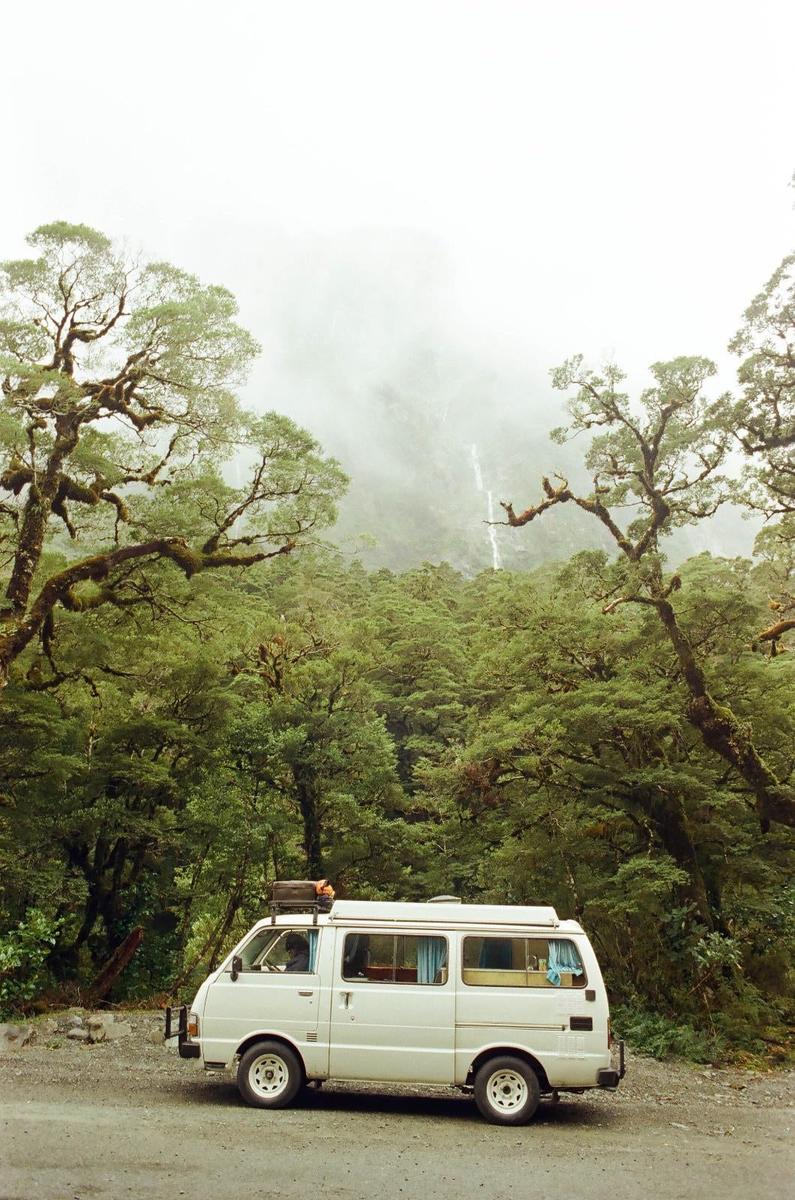 A white van is parked on a dirt road next to a dense forest with a waterfall visible in the misty background.