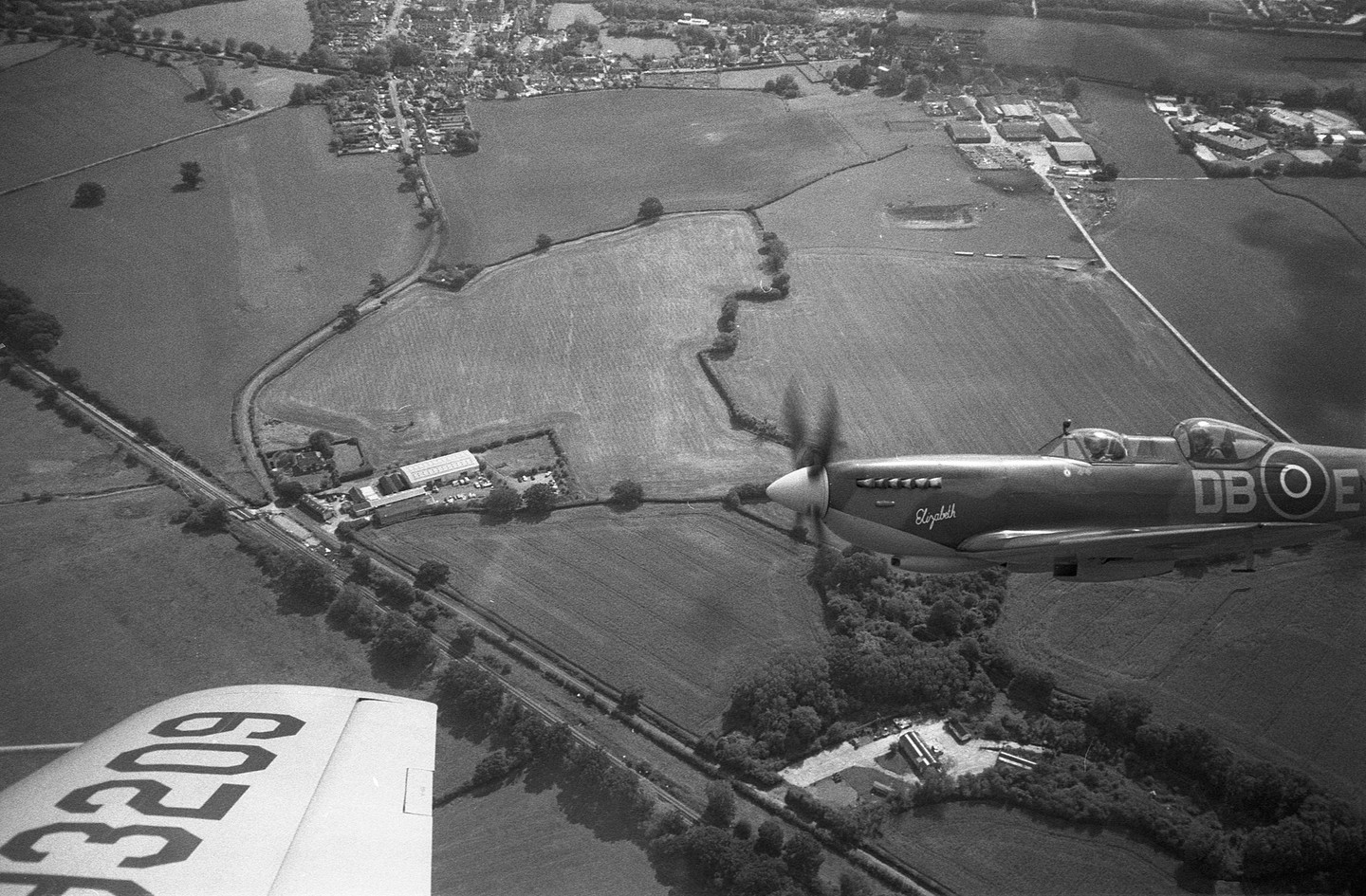 A Spitfire airplane flies over a rural landscape with fields and a small town visible in the distance.