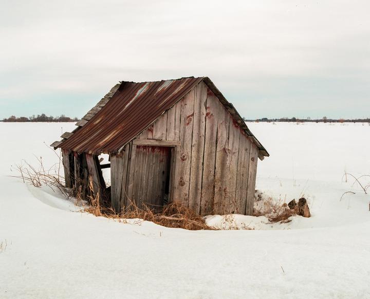 A weathered wooden shack with a rusty metal roof stands in a snow-covered field under a cloudy sky.