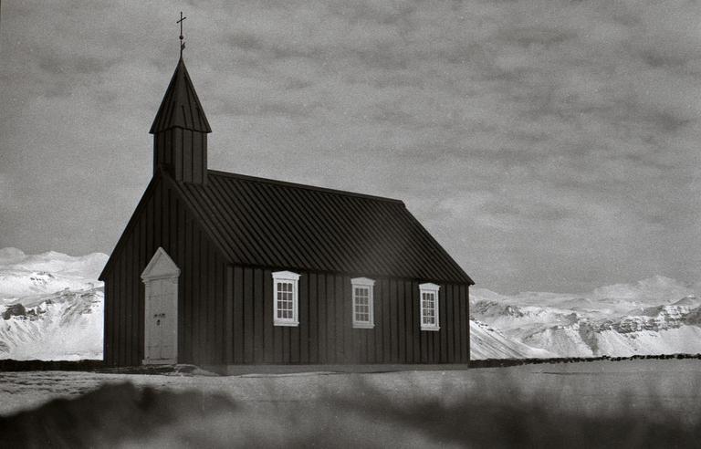 A black church with a steeple stands in a snowy landscape with mountains in the background.
