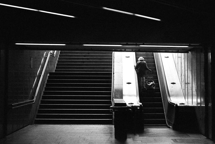 A woman with a shopping cart ascends an escalator next to a long staircase in a subway station.