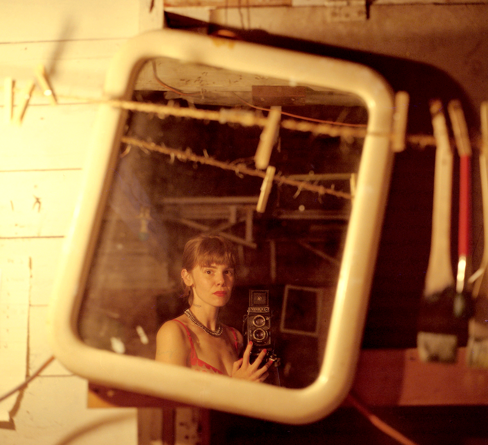A woman in a red top and a chain necklace holds a twin lens reflex camera while taking a self-portrait in a mirror.