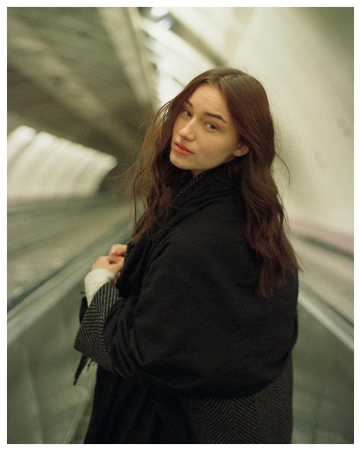 A woman with long brown hair looks over her shoulder while standing on an escalator.