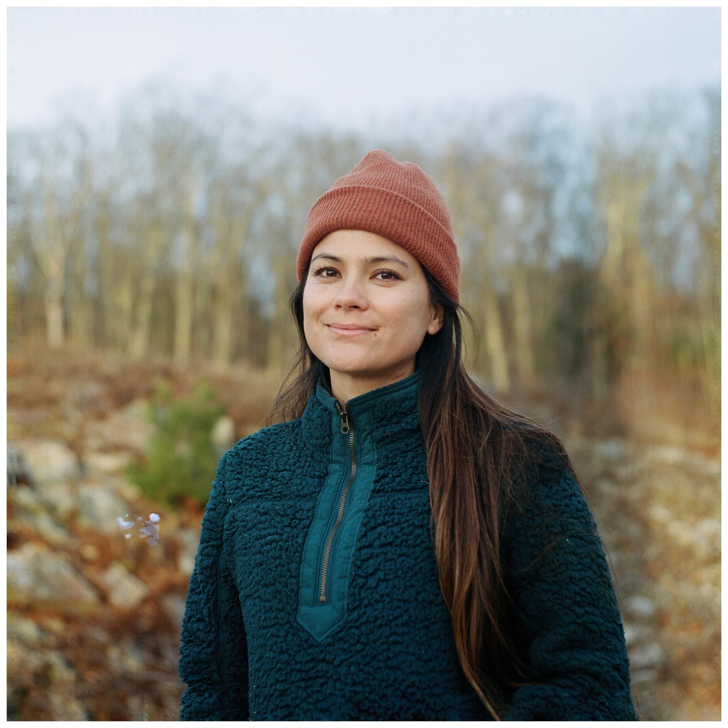 A woman wearing a beanie and a fleece jacket smiles at the camera with a forest in the background.