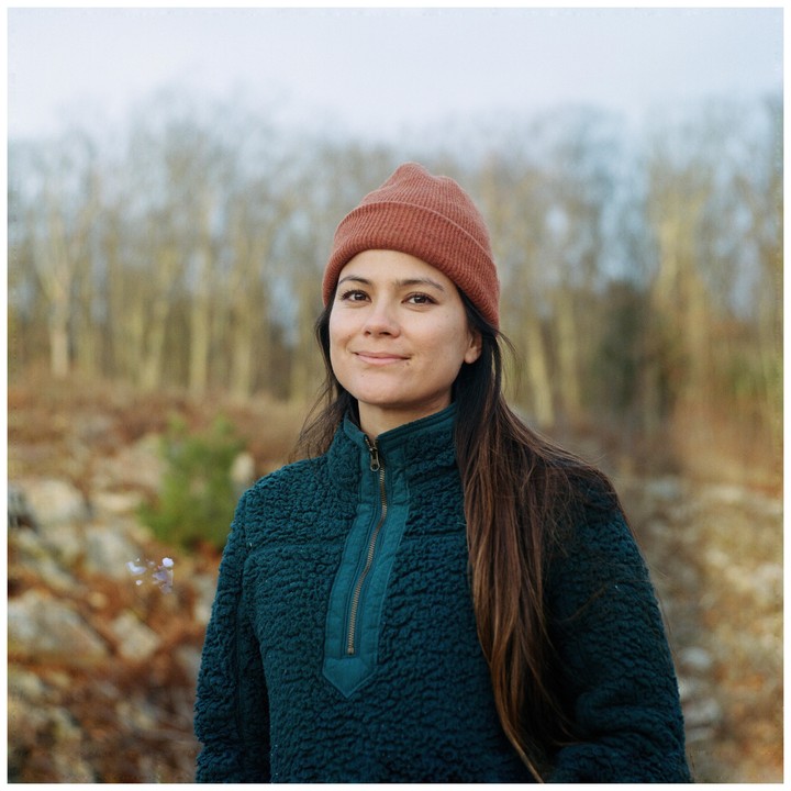 A woman wearing a beanie and a fleece jacket smiles at the camera with a forest in the background.