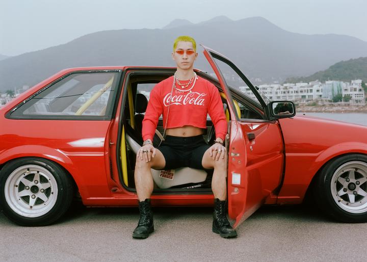 A man with yellow hair and red sunglasses sits in the open door of a red car with mountains and water in the background.
