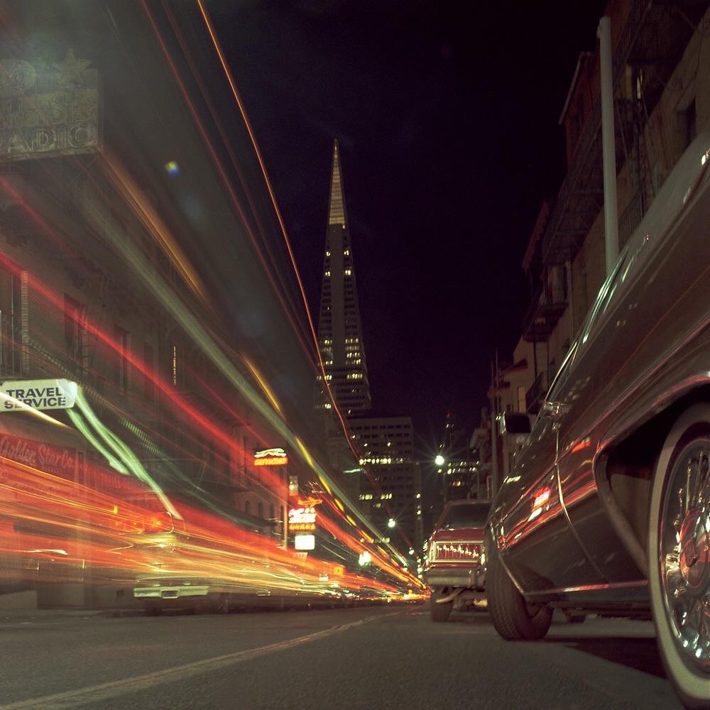 A street at night is filled with light trails from vehicles, with a tall, pointed building in the background and a parked car in the foreground.