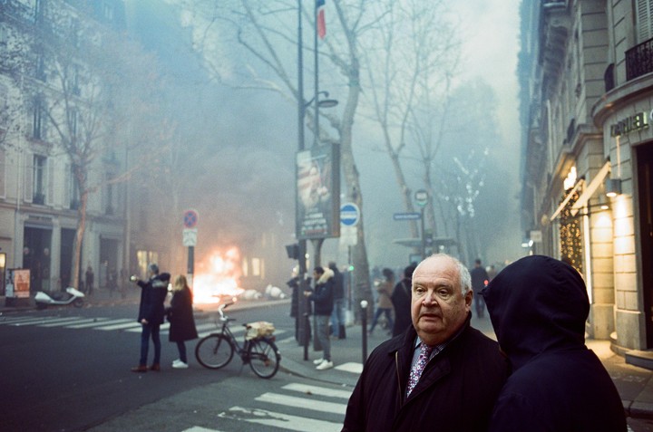 A man in a coat and tie looks forward while standing next to a person in a hoodie on a hazy street with a fire in the background.
